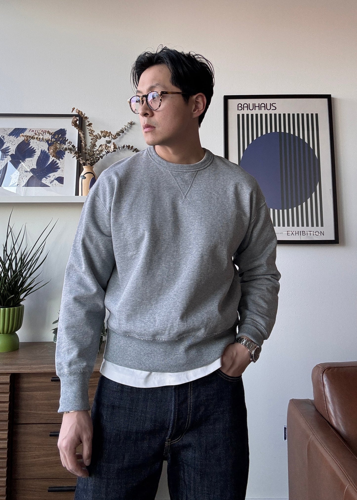 Light grey crewneck sweatshirt for men, shown layered over white tee in indoor setting, styled casually with raw denim. 