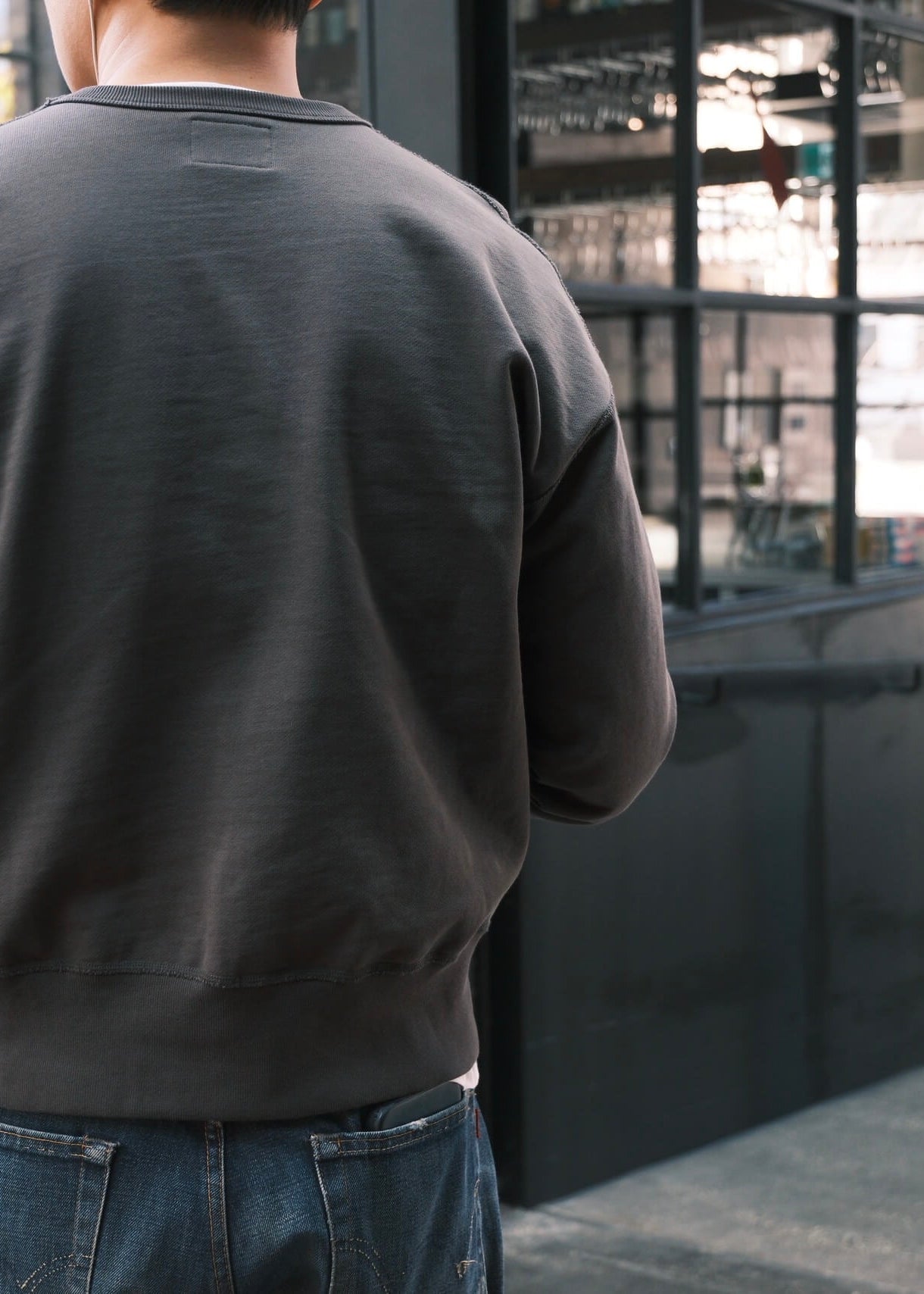 The back of a black crewneck sweatshirt styled casually with blue washed jeans, in an indoor setting. 